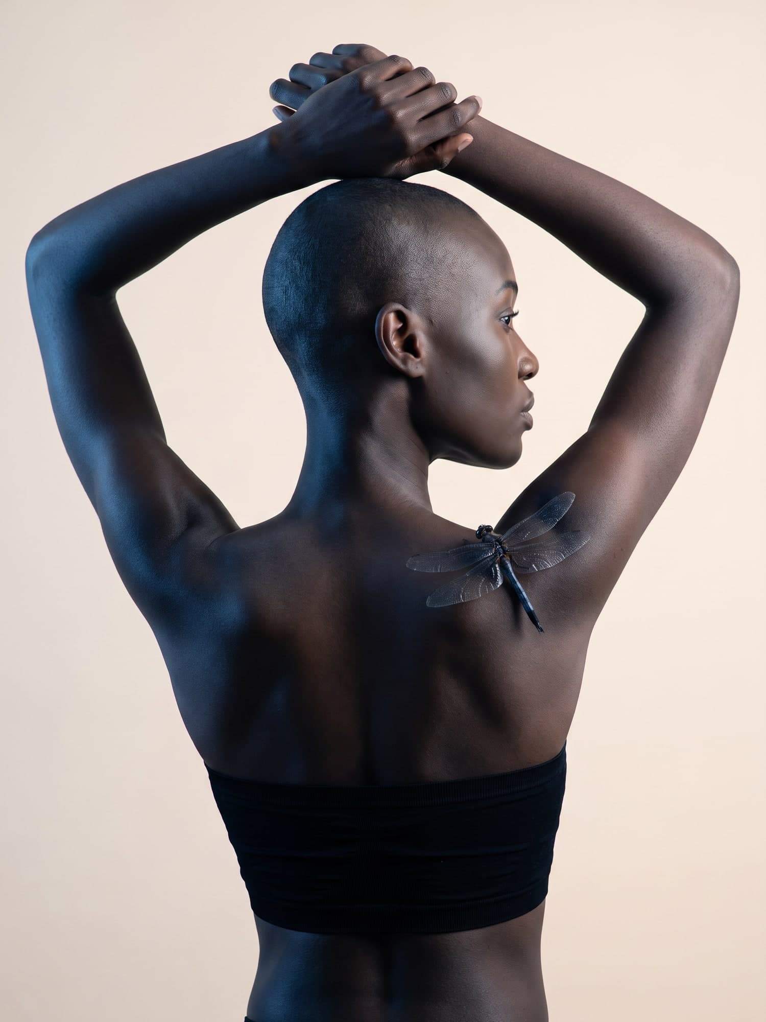 The model from behind, arms raised and hands clasped over her head, a dragonfly resting on her shoulder blade